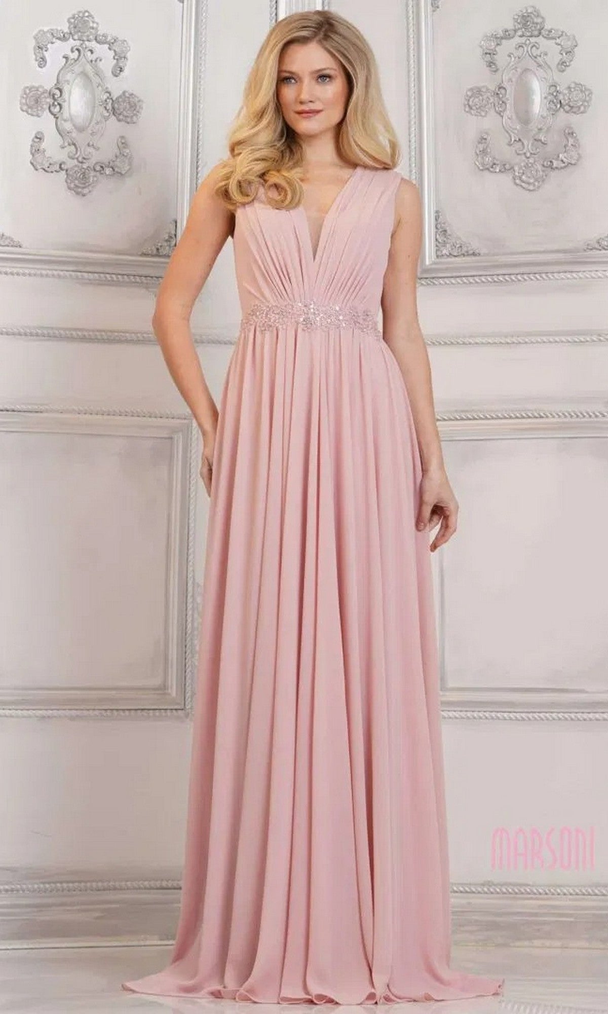A woman in a Marsoni MV1292 long formal dress in dusty rose, featuring a floor-length chiffon silhouette, sleeveless design, deep V-neck, ruched bodice, and beaded waist stands in an elegant light-colored room.